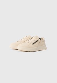 Beige leather sneakers with white laces, thick soles, and black side zippers, shown on a plain white background.