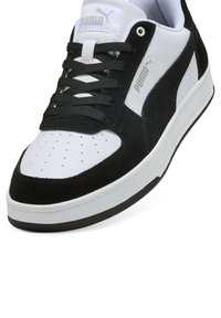 Zapatillas en blanco y negro con parte superior de ante y cuero, que presentan perforaciones, cordones planos y una suela de goma con un fondo texturizado.
