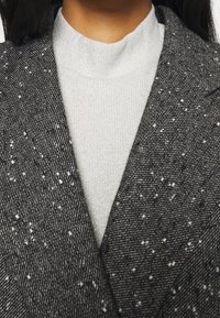 Gros plan sur une personne portant un blazer texturé noir et blanc tacheté par-dessus un pull à col montant gris clair.