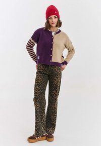 Pletený svetr v fialové a béžové barvě s proužkovanými rukávy, zapínání na knoflíky. Kalhoty s leopardím vzorem. Červená čepice a hnědé boty na platformě.