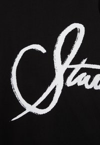 Texte blanc stylisé en cursive partiellement visible sur un fond en tissu noir, avec des bords texturés autour des lettres.