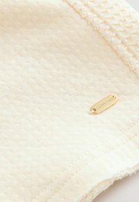 Cremefarbener Stoff mit strukturierter Oberfläche, versehen mit einem goldfarbigen Markenetikett. Sichtbare Nähte entlang der Kanten deuten auf qualitativ hochwertige Verarbeitung hin.