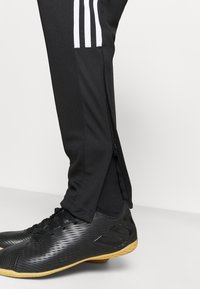 Black athletic pants with white stripes and zipper at the ankle, paired with black textured sports shoes featuring a tan rubber sole.
