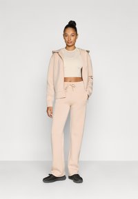 Ανοιχτό ροζ φούτερ με κουκούλα και αντίστοιχο παντελόνι φόρμας με κορδόνι στη μέση, σε συνδυασμό με μαύρα αθλητικά παπούτσια. Λεία υφή, άνετη εφαρμογή, ελάχιστες λεπτομέρειες.