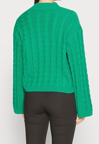Pull vert en maille avec un motif torsadé, col et ourlet côtelés, manches amples, assorti à un pantalon noir.