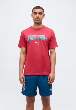 Fiatal férfi áll, piros "Portugália" Puma pólót és kék sport rövidnadrágot visel címerrel és Puma logóval, egyszínű háttér előtt.