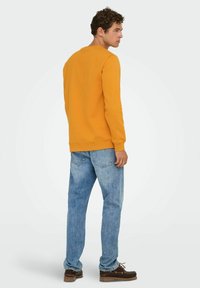 Geel sweatshirt met een ronde hals, geribbelde boorden en een relaxed fit, gecombineerd met lichtblauwe jeans en bruine schoenen.