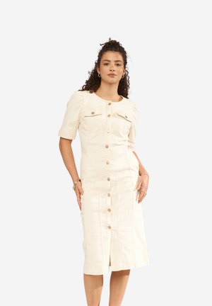 Žena s kudrnatými vlasy, oblečená v krémových šatech délky ke kolenům s knoflíky, krátkými rukávy a předními kapsami na prsou, stojící před jednoduchým pozadím.