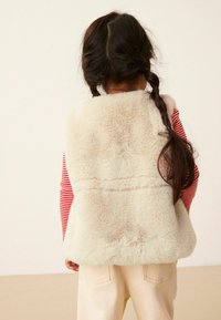 Gilet in finta pelliccia morbida e crema con colletto alto, indossato sopra una camicia a maniche lunghe rossa e bianca a righe, abbinato a pantaloni chiari.