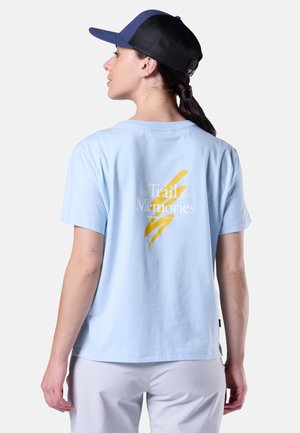 Vrouw draagt een lichtblauw T-shirt met "Trail Memories" en gele strepen op de achterkant, witte broek en een zwart-blauwe pet, kijkt naar links.