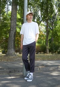 T-shirt blanc, pantalon foncé, baskets grises avec semelles blanches et casquette beige. Pris en extérieur dans un parc avec des arbres et un poteau.