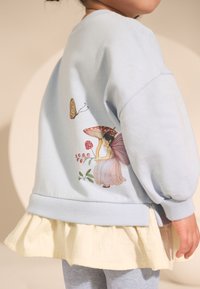 Jasnoniebieska bluza z balonowymi rękawami, zdobiona fantazyjną ilustracją wróżki. Warstwowa z falbanianym, jasnożółtym dolnym brzegiem.