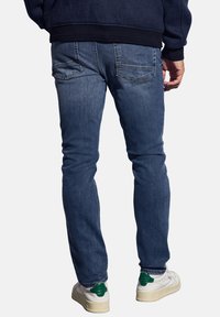 Hombre vistiendo jeans de mezclilla azul, zapatillas blancas con detalles verdes en el talón y una chaqueta oscura de color navy, de pie con las manos relajadas a los lados.