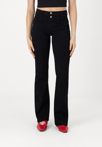 Jeans de campana negros con cintura alta, dos bolsillos frontales y cierre de botón. Combinados con zapatos planos rojos. Textura de denim suave.