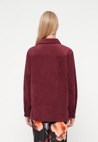 Bordowa kurtka z korduroy z kołnierzem, długimi rękawami i mankietami na guziki; ma wyraźny wzór i prosty krój.