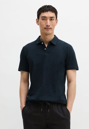 Man die een donkerblauw kortemouw poloshirt draagt met zwarte broek met trekkoord, staande voor een effen witte achtergrond.