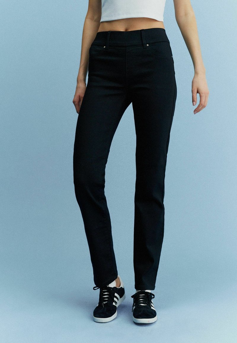 Schwarze Slim-Fit-Hosen mit glatter Textur, mit mittelhohem Bund und Taschen, kombiniert mit schwarzen Sneakers vor einem blauen Hintergrund.