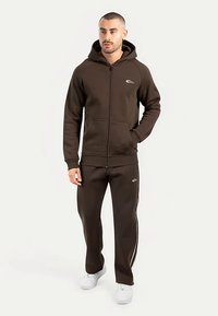 Sweat à capuche marron avec fermeture éclair, poches avant et logo ; assorti à un pantalon avec des bandes blanches sur les côtés. Les deux sont en tissu doux.
