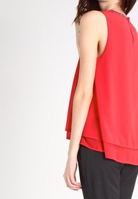 Top rosso senza maniche realizzato in tessuto leggero con un design a strati. Presenta una schiena con apertura a chiave e un accento decorativo sul collo.