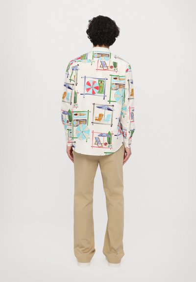 Bedrucktes Langarmhemd mit einem hellen Hintergrund und bunten Strandmotiven; kombiniert mit beigen Hosen.