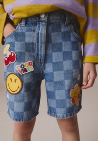 Short en jean avec un motif à carreaux, orné de patchs colorés : des cerises rouges, le texte "GOOD VIBES", un smiley et une fleur.