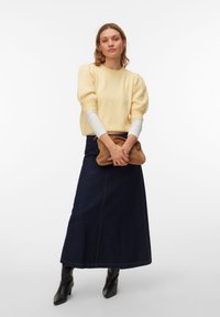 Femme portant un pull jaune à manches bouffantes par-dessus une longue chemise blanche, une jupe midi en denim foncé, des bottes noires, tenant une pochette marron.