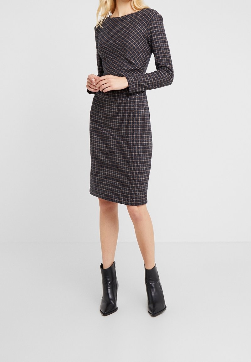 Donna che indossa un vestito aderente a maniche lunghe e lunghezza al ginocchio con un motivo a scacchi blu navy e marrone, abbinato a stivaletti neri con punta a punta.