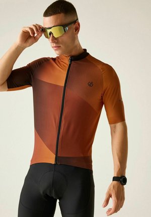 Man die geelgetinte sportzonnebril verstelt, draagt een bruin fietsshirt en zwarte gewatteerde fietsbroek tegen een effen achtergrond.