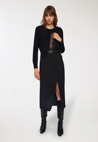 Cardigan nero con chiusura a bottoni, indossato sopra una blusa di pizzo nero trasparente, abbinato a una gonna nera con spacco laterale e stivali neri fino al ginocchio.