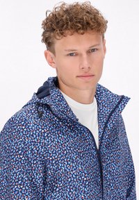 Giovane uomo con capelli ricci che indossa una giacca blu con motivo e zip sopra una camicia bianca, rivolto in avanti con un'espressione neutra.