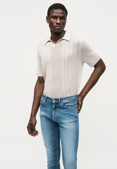 Άνδρας που φοράει ανοιχτό μπεζ πλεκτό πόλο με κοντά μανίκια και μπλε τζιν, στέκεται με το ένα χέρι στη μέση μπροστά από απλό φόντο.