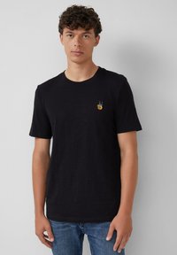 Jonge man met krullend haar, gekleed in een zwart T-shirt met een klein kleurrijk vredesteken op de borst en blauwe spijkerbroek.