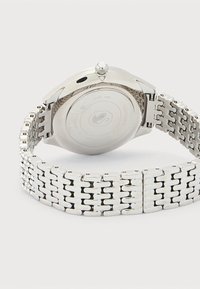 Ασημένιο ρολόι χειρός από ανοξείδωτο ατσάλι με συνδεδεμένο μπρασελέ, ορατό κορωνάκι και κλειστή αγκράφα, εμφανίζεται από πίσω σε απλό ανοιχτό φόντο.