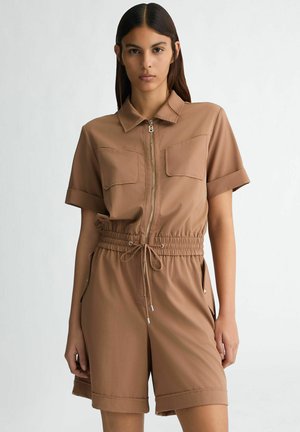 Tan jumpsuit featuring short sleeves, a zip front, two chest pockets, elastic waist, and rolled cuffs. The fabric appears smooth and lightweight.