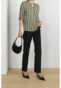 Femme portant un blouse rayé vert olive et blanc, un pantalon noir, des escarpins noirs à brides, tenant un petit sac à main noir, debout à l'intérieur.