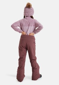 Lila Fleece-Pullover mit geometrischem Muster und hohem Kragen, kombiniert mit burgunderfarbenen Schneehosen. Eine Strickmütze mit Pompon rundet den Look ab.