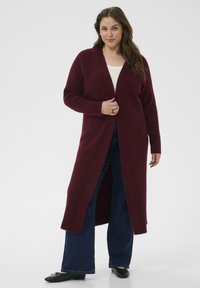 Cardigan lungo bordeaux con texture a coste, davanti aperto e maniche lunghe, abbinato a jeans blu a gamba larga e ballerine nere.