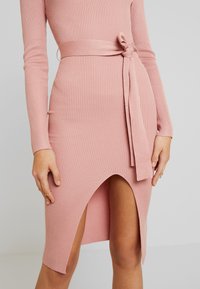 Rosa ribbad klänning med en figurn ära silhuett, långa ärmar och ett knytbälte. Har en hög slits på vänster sida för extra detalj.