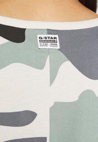 T-paita, jossa on kamouflagekuvio vihreillä, harmailla ja kermavärisillä sävyillä. Ominaisuuksiin kuuluu valkoinen kaulus ja etiketti, jossa lukee "G-Star Originals RAW Denim."