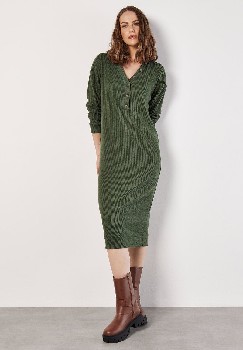 Langes grünes geripptes Kleid mit V-Ausschnitt, Knopfdetails und langen Ärmeln. Kombiniert mit braunen Ankle Boots mit einer klobigen Sohle.