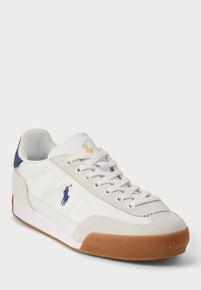 Fehér, alacsony szárú sneaker bézs velúr részletekkel, tengerészkék és arany Polo logóval, kék sarokrésszel, fehér fűzővel és gumírozott talppal.