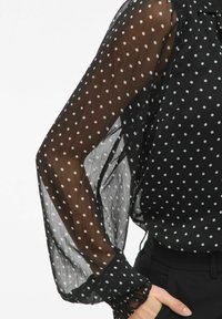 Zwarte transparante blouse met witte polkadots, lange mouwen en opgepofte manchetten. De stof is lichtgewicht met een licht gekreukte textuur.