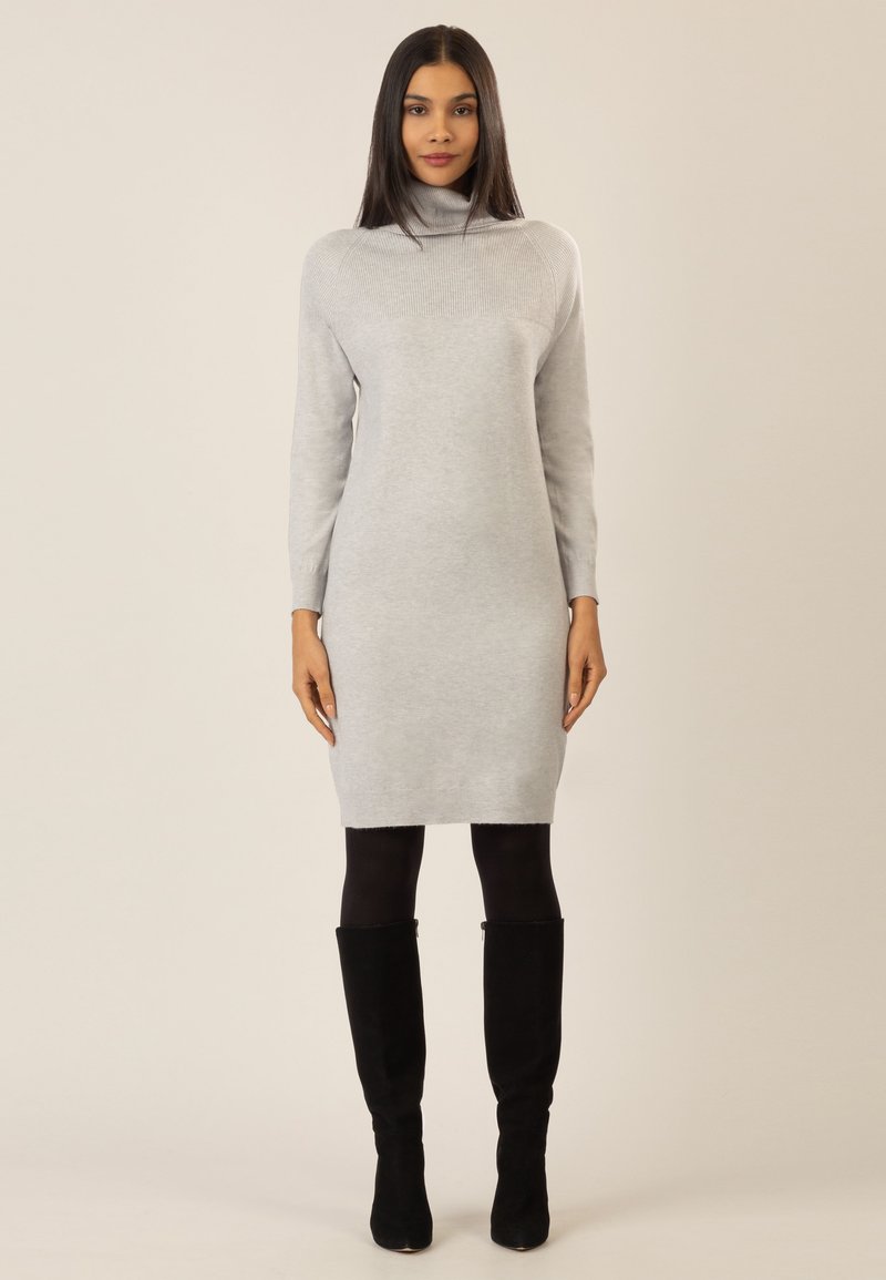Graues Strickkleid mit hohem Kragen, langen Ärmeln und taillierter Silhouette, kombiniert mit schwarzen Overknee-Stiefeln. Glatte Textur.