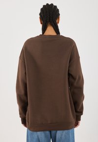 Person med flettehår iført en løs brun hettegenser og lyseblå jeans, vendt bort fra en ensfarget lys bakgrunn.