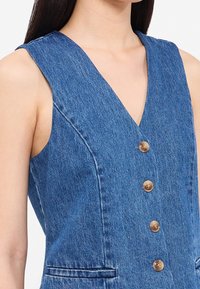 Jeansklänning utan ärmar med djup V-ringning, mässingsknappar och fickor. Tyget har en lätt strukturerad yta i mellanblått.