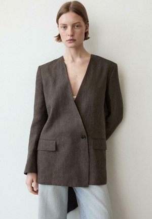 Junge Person mit kurzen Haaren trägt einen übergroßen braunen Blazer über hellblauen Jeans, steht vor einer schlichten hellen Wand und schaut geradeaus.