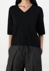 Suéter negro de manga corta y escote en V en un tejido suave, combinado con pantalones oscuros de cintura alta y textura, que presentan cordón y detalles de pliegues.