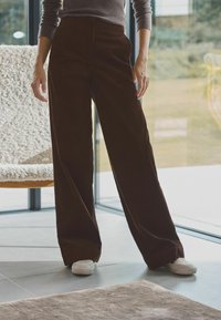 Brune kordfløjls bredbenede bukser med høj talje, suppleret af hvide sneakers, sat mod en lys, moderne indretning.