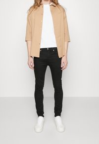 Persona vestida con una camisa beige de botones sobre una camiseta blanca, jeans ajustados negros y zapatillas blancas, de pie contra un fondo simple.
