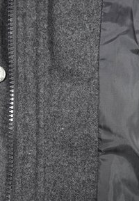 Grå jakke med uldlignende tekstur på den ene side og glat, sort nylon på den anden, med lynlås og metaltryklåse som detaljer.